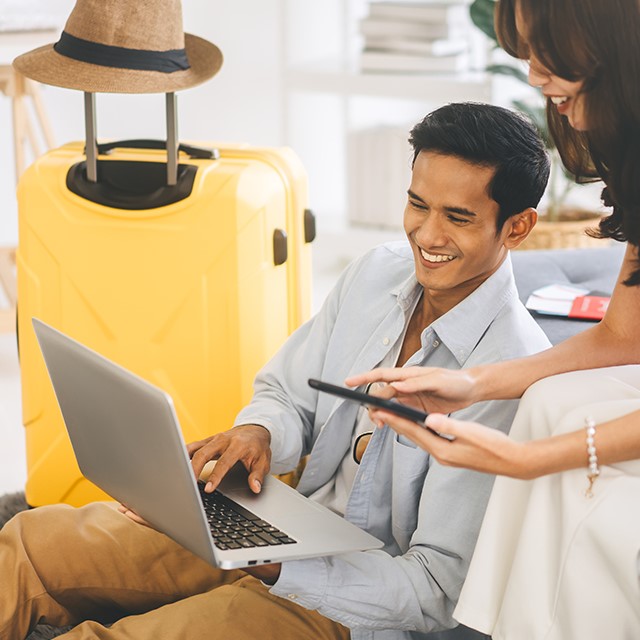 Mann hält Laptop und liest mit Frau Reisebewertungen, neben Ihnen steht ein gelber CHECK.IN Koffer mit Strohhut auf dem Griff