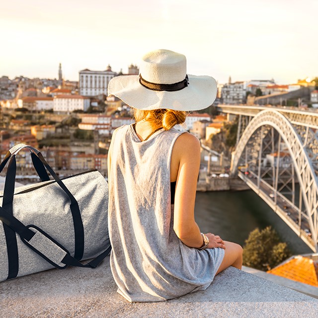 Frau mit Sonnenhut und Reisetasche sitzt auf Mauer und blickt über die Stadt